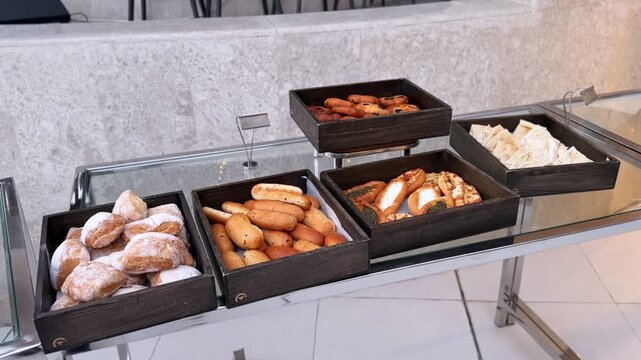 Breakfast buffet table featuring assorted fresh bread rolls, powdered donuts, savory stuffed pastries, and sliced flatbread in rustic wooden trays on glass stand indoors.