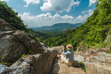 Young woman sitting on rocky viewpoint in tropical mountains taking photo of green valley and...
