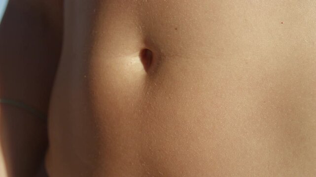 CLOSE UP, DOF: The belly and navel of a fit young woman in bikini illuminated by the sun. Skin is warm and smooth, showing the soft texture of a summer tan during a relaxing sunny day at the shore.