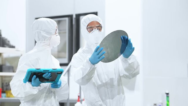 Male and female workers in protective clothing inspecting silicon wafers with tablets in a semiconductor factory