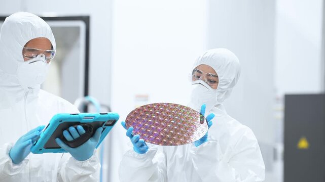 半導体工場でタブレットを持ってシリコンウエハーを検査する防護服を着た男女作業員