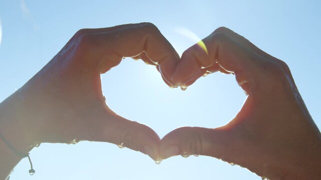 CLOSE UP, LENS FLARE: Wet hands come together to form a heart shape, framing the summer sun. Tiny droplets of water glisten on the skin, capturing a moment of pure happiness and love for the ocean.