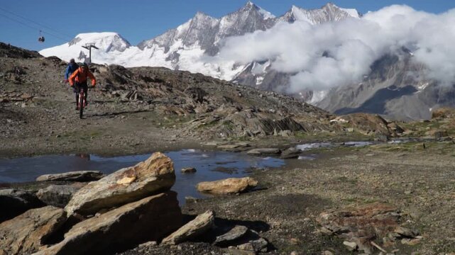 Two mountain bikers riding downhill and crossing a small alpine river in Saas-Fee, Swiss Alps, with dramatic 4000m peaks in the background. Dynamic splash, high alpine scenery, cinematic adventure ene