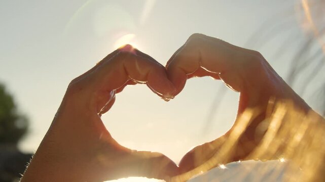 CLOSE UP, LENS FLARE: Young female hands form a heart shape against the glowing summer sun. Tiny droplets of water sparkle on the skin. Moment of pure happiness on the beach and love for the sea.
