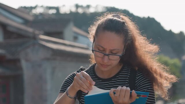 Woman sketches architecture in notebook on sunny street, focused student vibe with pen and glasses, heritage facades and quiet urban courtyard provide inspiration for study and art