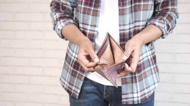 A man in a plaid shirt and jeans holds an empty wallet with no money, representing financial hardship and economic crisis