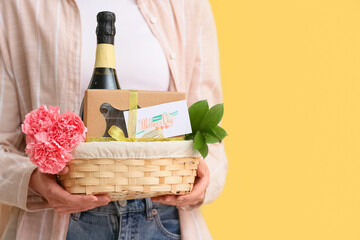 Young woman holding basket with carnations, champagne bottle and chocolate candies for Mother's Day on yellow background © Pixel-Shot