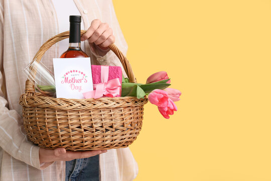 Young woman holding basket with wine bottle, tulips and gift box for Mother's Day on yellow background