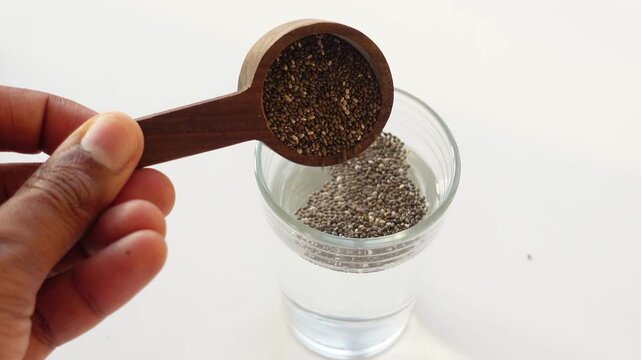 Wooden spoon pouring chia seeds into a glass of clear water on white background, healthy lifestyle choice