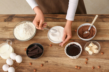 Obraz premium Woman adding spoon of sugar into bowl with chocolate for preparing brownie at table in kitchen. Closeup