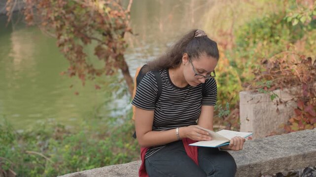 Writer by river. Note taker seated near flowing stream. Individual jotting ideas beside tranquil riverbank. Person documenting thoughts overlooking calm water with autumn leaves falling