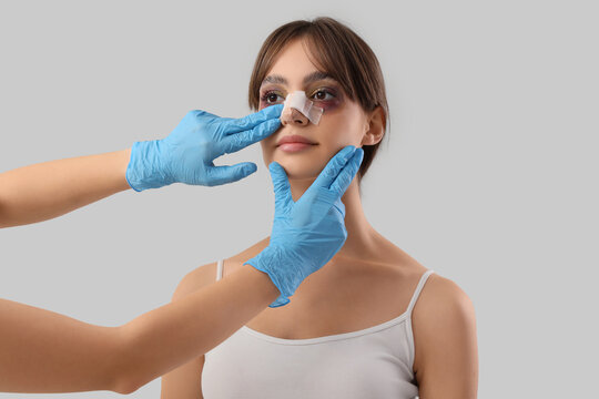 Surgeon hands touching woman's bandaged nose after plastic surgery on grey background