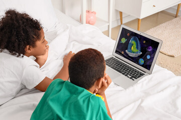 Fototapeta premium Cute African-American children watching cartoons on laptop in bedroom at home