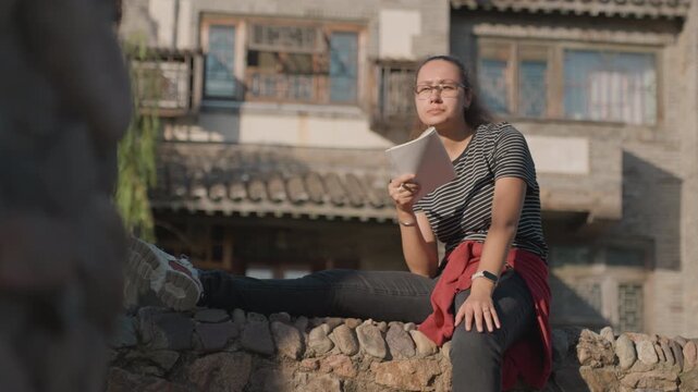Young woman reading letter on stone wall in historic quarter, striped shirt and glasses, red jacket tied at waist, sunlight casting warm glow, quiet courtyard ambience, handheld paper, subtle smile,