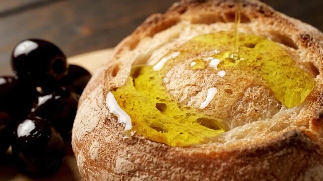 Olive oil pours onto bread with dark olives nearby, wooden background, focus on texture