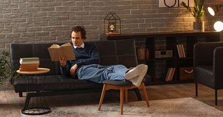 Fototapeta premium Young man reading book on sofa at home late in evening