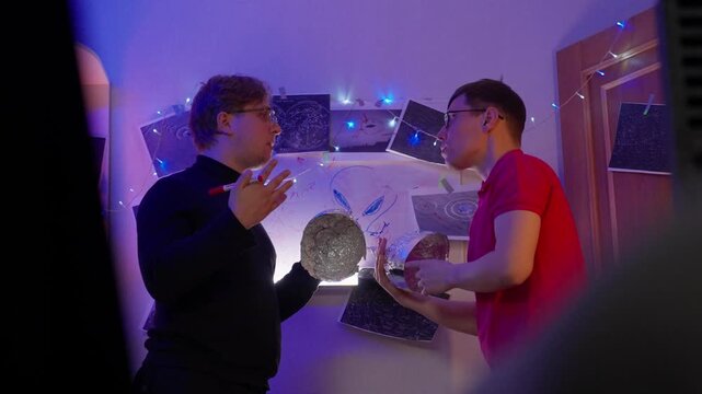 Two White Men Wearing Foil Hats. Amateur Ufologists Examine Reflective Sphere Under Purple Led Lights In Cramped Studio, Pointing At Space Posters And Star Maps, Animated Debate About Alien Signal