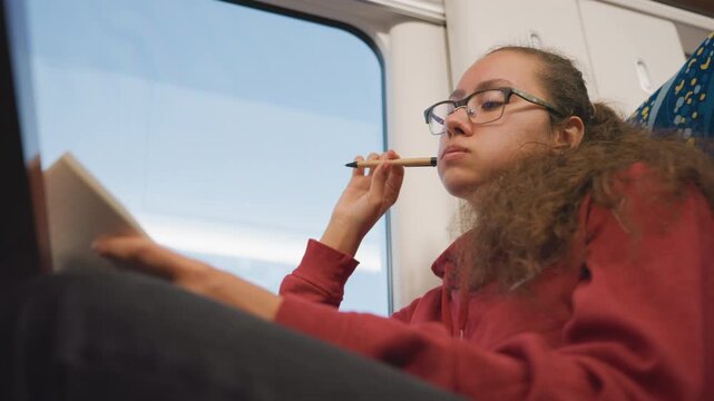 Young woman rehearsing lines aloud on train, notebook held, expressive gesture, vocal practice, glasses and intent gaze, passing landscape outside window, performance prep, monologue practice, focused