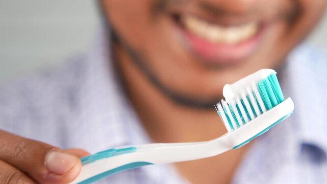 A man&rsquo;s hand holds a toothbrush with toothpaste against a blurred background, a smile is visible, highlighting good oral hygiene