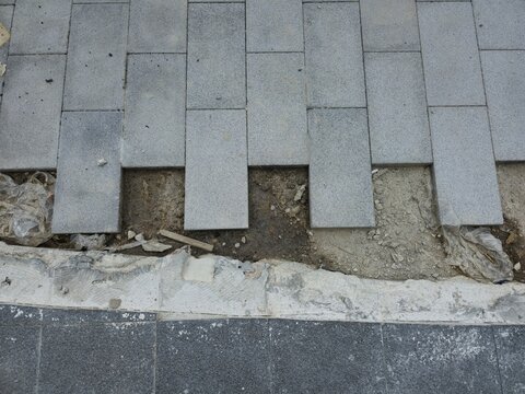 A cracked sidewalk with a few missing bricks. The sidewalk is made of gray bricks and is located next to a street