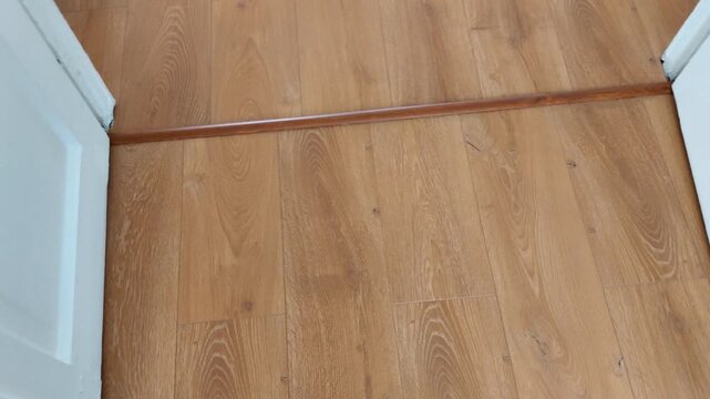 Light brown wooden floor planks meet at a threshold between rooms, with white doors slightly open, highlighting everyday home detail and architectural transition