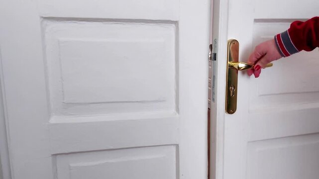 A hand in a red sleeve opens classic white double doors with a gold handle, capturing the everyday action of entry and exit with a simple, clear household detail