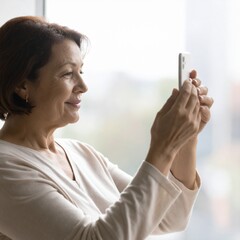 明るい朝の室内、窓辺で撮影されたフォトリアルな医療サービス広告向けスチル写真。
50代半ばの女性がスマートフォンを胸の高さで持ち、画面を穏やかに見つめている。