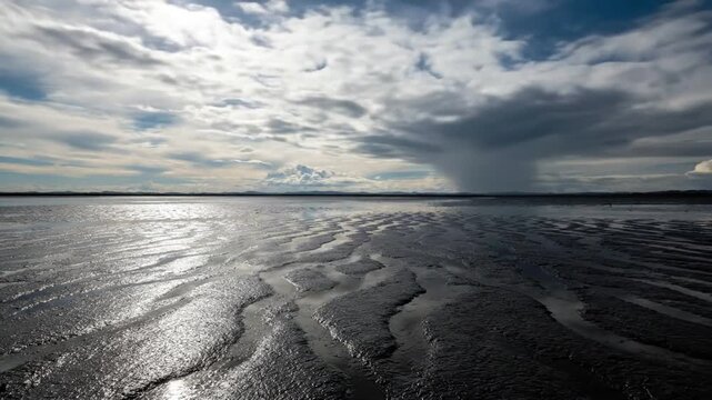 Dynamic Cloud Patterns Emphasize the rapid transformation and movement of various cloud types (cirrus, cumulus, nimbostratus) as a time-lapse, both in the sky and mirrored in the mud.