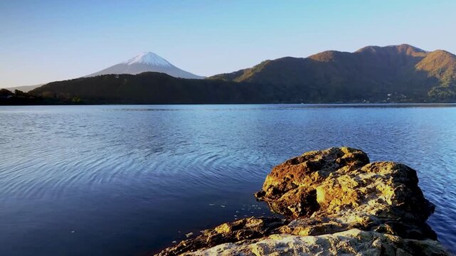 Stunning timelapse of Mount Fuji at dawn with calm lake reflection in the foreground and volcanic rocks on the shore. Morning light gradually illuminates the iconic snow capped peak against a clear bl
