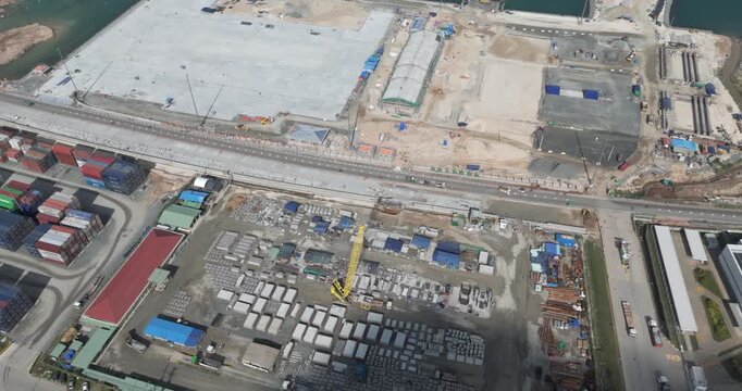 Top down aerial view of the Sihanoukville port expansion, featuring a yellow crawler crane, road construction, stacked shipping containers, and vast new concrete terminal foundations.