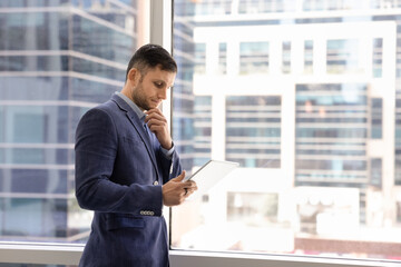 Fototapeta premium Focused businessman in suit stands by panoramic office window, thoughtfully analyze data or read information on tablet, ponder on idea, solution, make important decision. Analytical thinking, planning