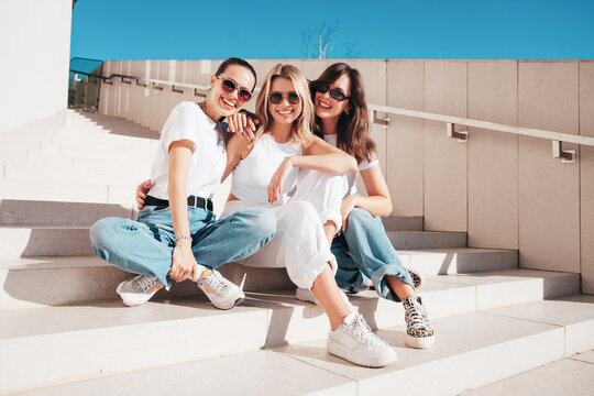 Three young beautiful smiling hipster female in trendy summer t-shirt  and jeans clothes. Sexy carefree women posing in street. Positive models having fun, sit at stairs. In sunny day, sunglasses
