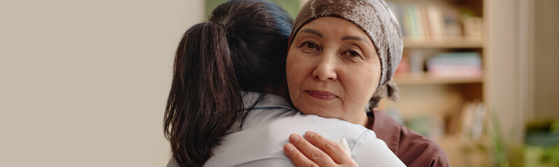 Header portrait of middle aged Asian woman wearing headscarf hugging young adult woman, looking...