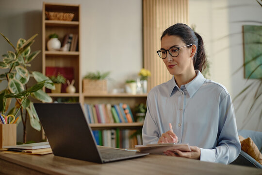 Caucasian young adult woman sitting at desk using laptop, wearing eyeglasses, focusing on online psychology work or virtual therapy session in modern office setting