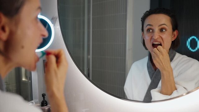 Woman in a bathrobe using a dental floss pick in front of a bathroom mirror during her morning hygiene routine. She carefully cleans between her teeth, maintaining oral health and personal care. The