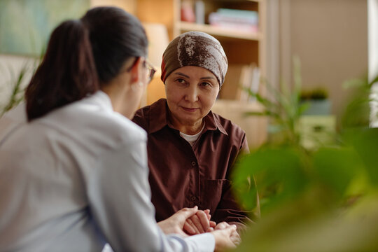 Middle aged Asian woman wearing headscarf sitting with young adult woman holding hands, engaging in supportive conversation during psychological counseling session