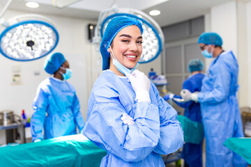 Successful female surgeon smiling with mask down in operating room. Confident medical professional with crossed arms after surgery. Healthcare and medicine concept. Team of doctors in background. © Graphicroyalty
