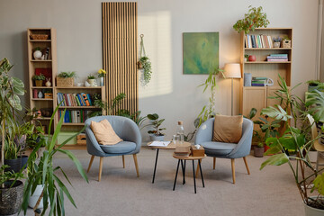 Modern psychology office interior featuring two empty armchairs facing small table with books, glass water carafe, clipboard and pen, surrounded by numerous green houseplants and bookshelves