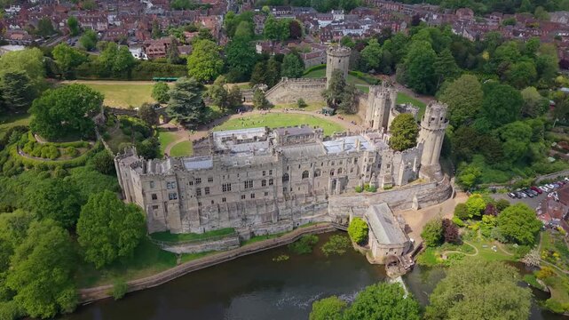 Aerial drone pullback from above revealing Warwick Castle Dungeon and the surrounding historic town of Warwick on a bright sunny day, showcasing medieval architecture and green landscape.