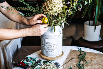 Hands place small green fruit into rustic floral arrangement with hydrangea and leafy branches in...