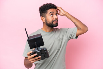 Brazilian man holding a drone remote control isolated on pink background having doubts and with...