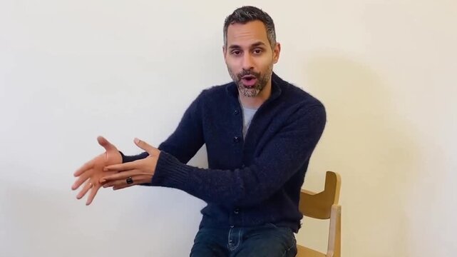 Man speaking seated by empty wall. Young Hispanic businessman engaged in business meeting or discussion seated at desk in modern office, presents sales result and progress, project updates, proposing 