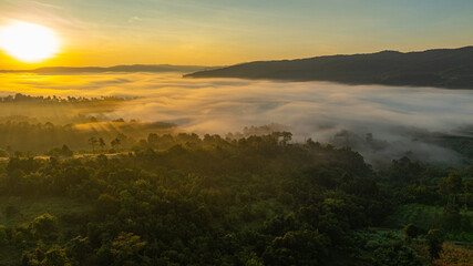 Aerial sunrise casts warm golden light over a mist covered forest and rolling hills, creating a serene and atmospheric landscape scene. Ideal for themes of nature, tranquility, and new beginnings. © Narong Niemhom