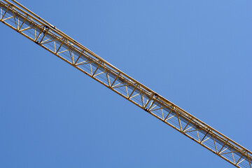A long yellow metal truss of a construction crane jib stretches diagonally across a clear blue sky background under bright sunlight