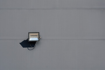 A modern black LED floodlight is mounted on a gray corrugated metal building facade casting a sharp shadow under bright daylight