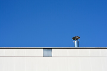 A stainless steel chimney pipe with a rain cap stands on the edge of a flat white industrial building roof against a clear blue sky