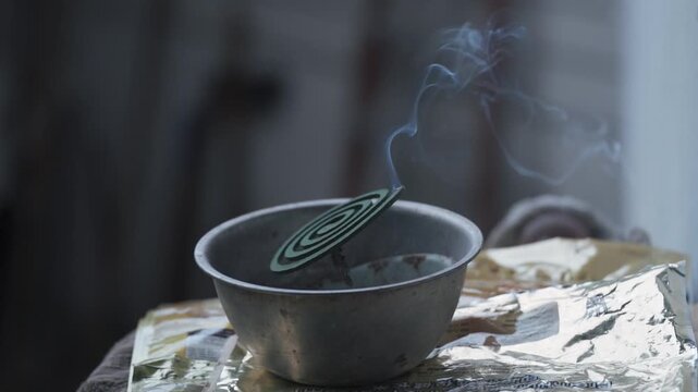 A close-up of a green spiral mosquito coil burning inside a rustic metal bowl, with a steady wisp of smoke rising, commonly used as an insect repellent in tropical climates.
