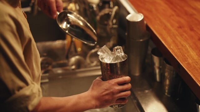 A close-up of a professional bartender using a stainless steel scoop to fill a cocktail mixing tin with large, clear ice cubes at a trendy dimly lit bar.