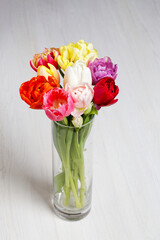 Fototapeta premium Vibrant spring tulips bouquet in cylindrical glass vase on white wooden table. Copy space. Minimalist floral decor for Mother's Day, greeting card or banner design.