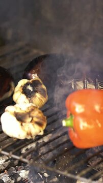 Close-up of fresh red pepper, garlic and eggplant roasting on a grill with smoke and open flames, preparing the traditional Mediterranean Escalivada.